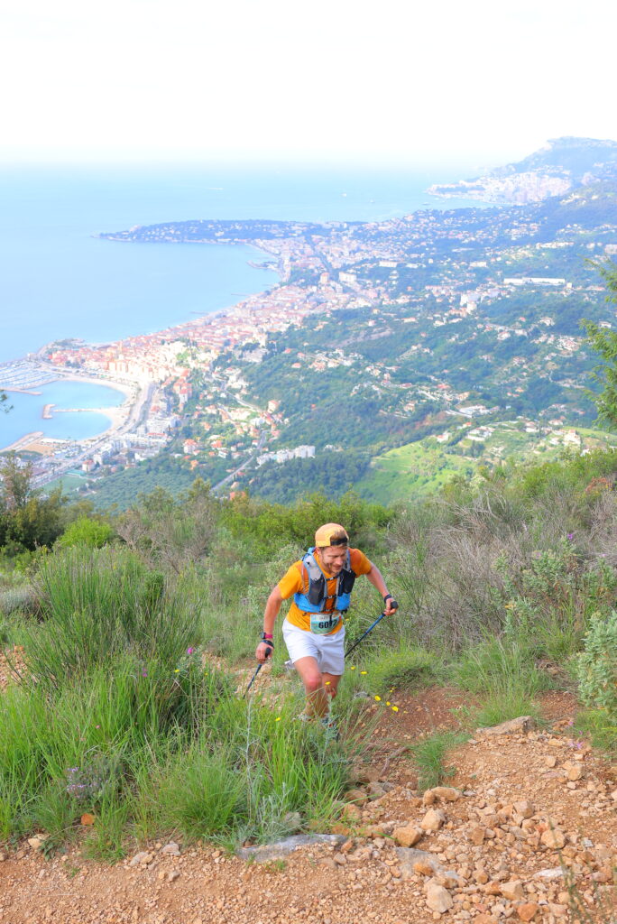 coureur trail en train de gravir le Roc Orméa, ligne cotière de Menton à Monaco visible en arrière plan